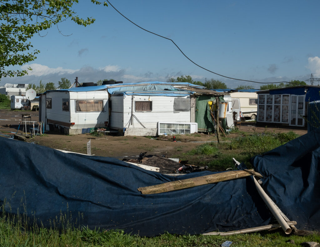 au premier plan il y a une palissade en toile affaissée et des bouts de bois posés dessus. Au second plan on voit plusieurs caravanes regroupées dont certaines sont collées les unes aux autres.