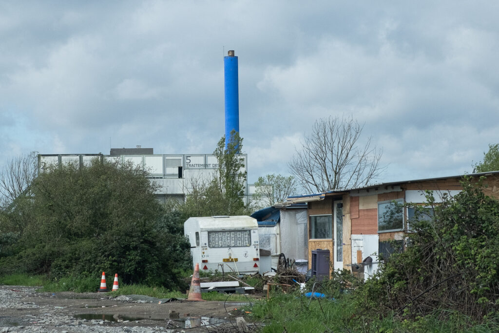 Au premier plan un sol caillouteux avec des flaques. Au second plan des caravanes et cabanes. En arrière plan un hangar de la déchèterie et une grande cheminée d'incinération des déchets.