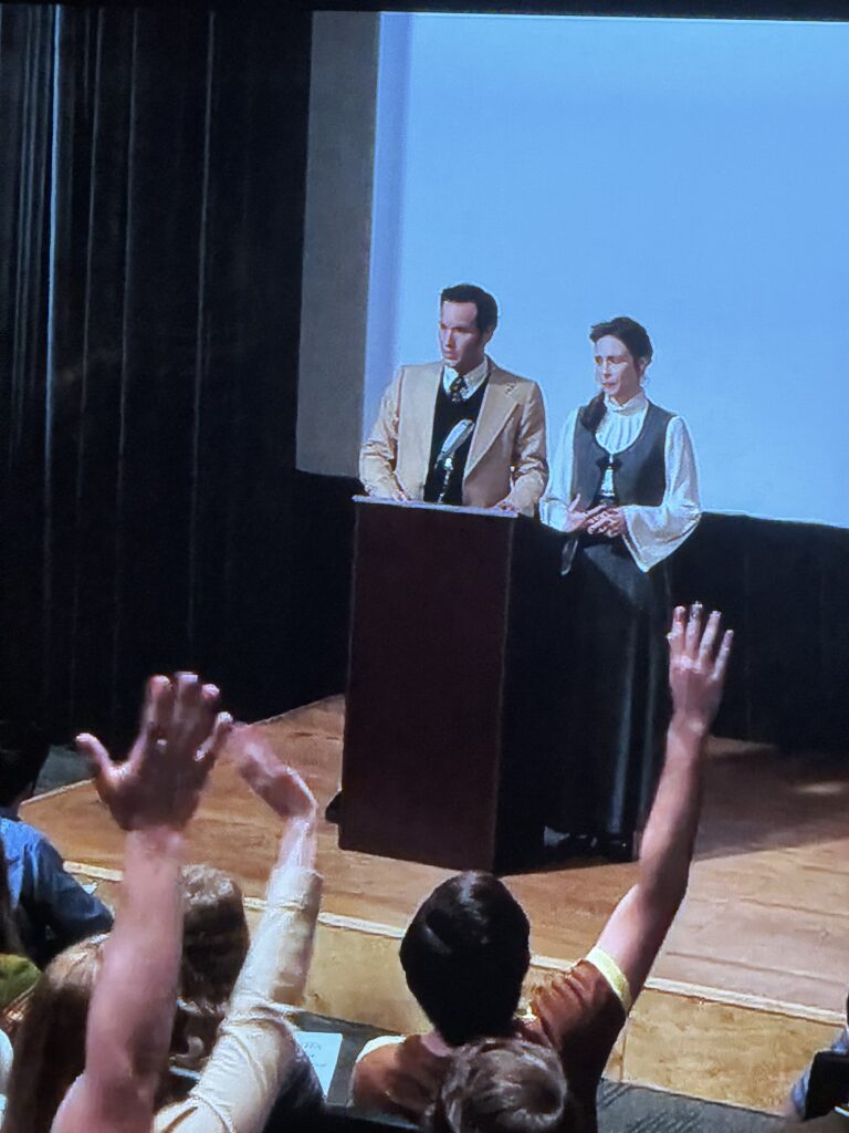 ce plan du film montre les époux Warren donner une conférence, de multiples étudiants lèvent la main