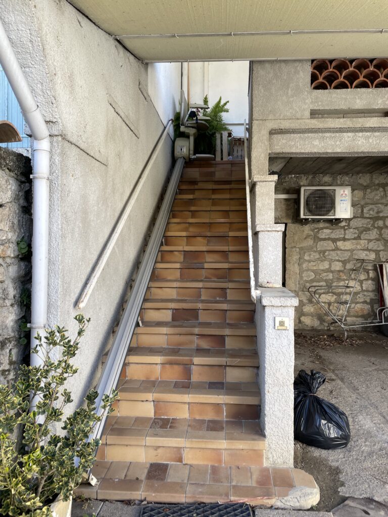 un escalier extérieur menant à une maison, une rampe a été installée permettant de monter les escaliers via une chaise