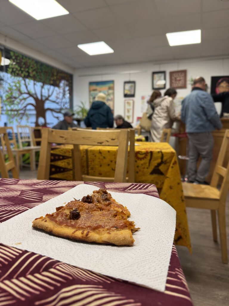 plusieurs petites tables couvertes de nappes sont disposés au sein de la salle du café associatif. près du bar au fond on entrevoit les silhouettes des participantes et participants. Au premier plan une pizza est posée sur une table