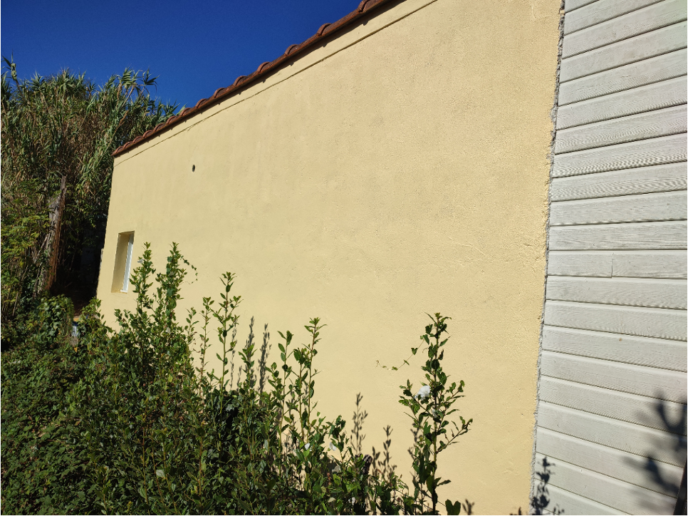 au premier plan un grand mur  peint au jaune pale qui se termine sur un volet en alluminium fermé. De grandes herbes poussent le long du mur. 