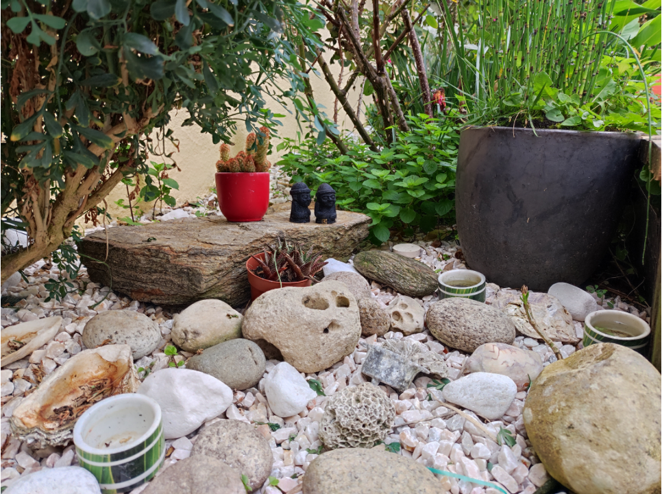 au premier plan sur des galets blancs sont posés des pierres beige et blanches  de différentes tailles. Au premier plan on aperçoit une bougie presque complètement consumé dans un pot en céramique coloré. au second plan il y a un cactus en pot et deux  statuettes représentants des silhouettes rondes. 
