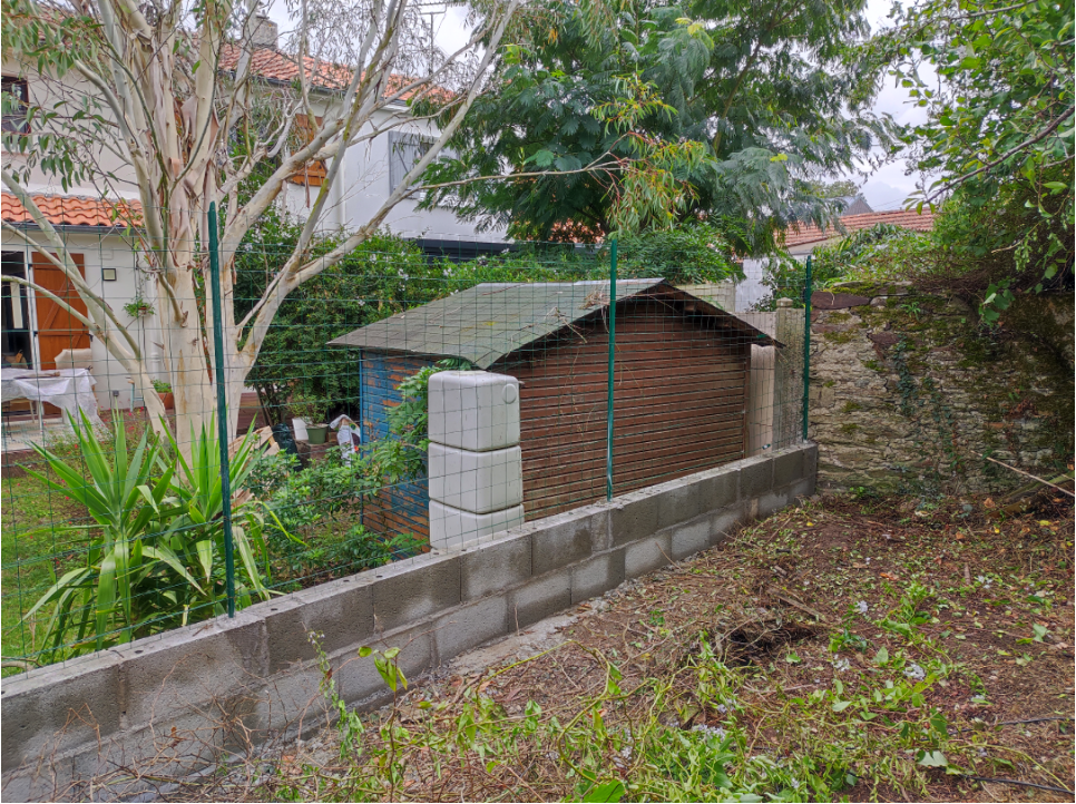 au premier plan on voit de la terre et un muret fraichement monté. Derrière le muret il y a un autre jardin avec une cabane de jardin et un peut plus loin un immeuble.