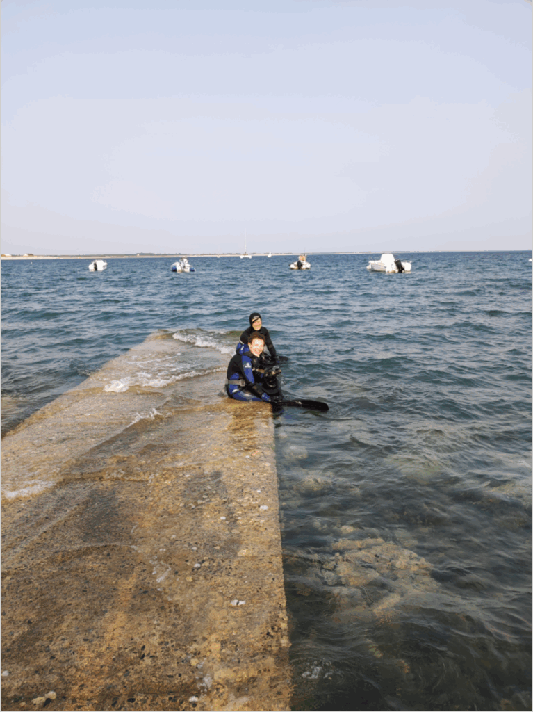 Un homme et une femme en combinaisons de plongée sont assis les pieds dans l'eau sur la pente en béton qui permet de mettre les bateaux à la mer. Au long des petits bateaux sont au mouillage